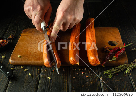 Slicing sausage with a knife in the hand of a chef on a kitchen board before preparing dinner. Low key concept for serving meat food Slicing sausage with a knife in the hand of a chef on a kitchen board before preparing dinner. Low key concept for serving meat food 112500680