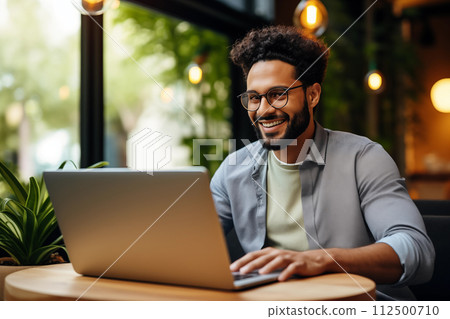 Young, happy male enjoying remote work from a warm, well-lit coffee shop 112500710