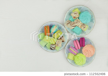 Various desserts. Sweet desserts in plates for holiday. Macaroons, marshmallows and zephyrs on a white background. Top view. Various desserts. Sweet desserts in plates for holiday. Macaroons, marshmallows and zephyrs on a white background. Top view. 112502406