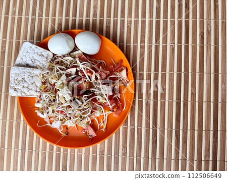 Salad of fresh vegetables and micro-greens. Tomatoes, eggs, bread rolls, onions, apples, mound of sprouted mung beans in an orange bowl on bamboo background. Concept of diet, vegan, healthy products. 112506649