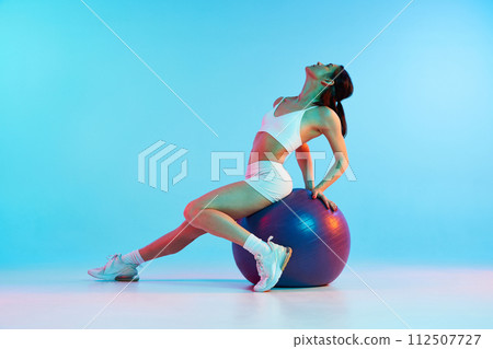 Slim, sportive young woman in white sportswear sitting on fitness ball, training against blue studio background in neon light Slim, sportive young woman in white sportswear sitting on fitness ball, training against blue studio background in neon light 112507727
