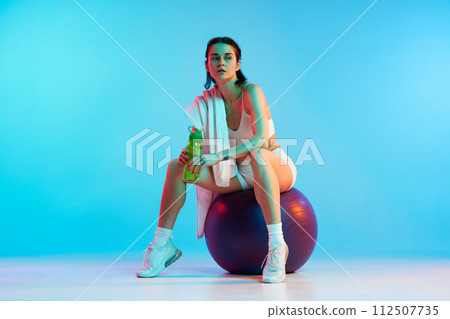 Young woman in sportswear sitting on fitness ball with water and towel against blue studio background in neon light. After training 112507735