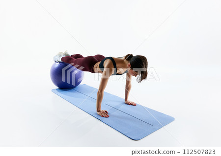 Sportive young woman with fit body training, standing in plank position on fitness ball isolated over white studio background 112507823