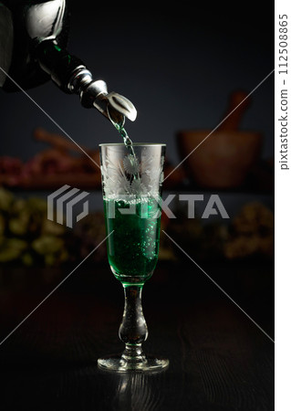 Green herbal liquor is poured from a vintage bottle into a glass. Green herbal liquor is poured from a vintage bottle into a glass. 112508865