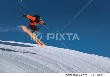 A skier in an orange jacket and a black helmet makes a jump on a sunny day against a clear sky. Copy space for your information 112509300