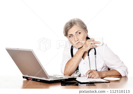 Serious old woman doctor sitting at table with computer Serious old woman doctor sitting at table with computer 112510113