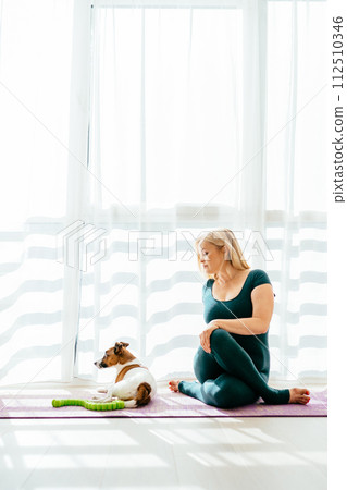Vertical shot of middle age blond 55s woman in sportswear channels serious expression while sitting on yoga mat with her dog in Livingroom. Person's balance of a healthy, indoor sport lifestyle. 112510346