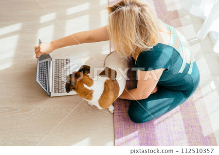 Healthy mature 55s woman training at home, watching online videos on laptop. Active dog Jack Russell wants to play, is in the way for her. Female connecting online yoga classes practicing exercise 112510357