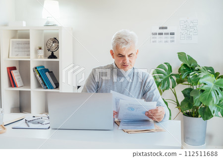 Middle aged businessman reading paper documents and using laptop while working at home office. Confident, experienced senior male professional. Small business entrepreneur manage business paperwork 112511688