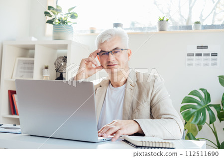Portrait of smiling grey hair middle-aged man working on laptop while sitting at his work place in office. Confident, experienced senior male professional. Small business entrepreneur manage business. 112511690