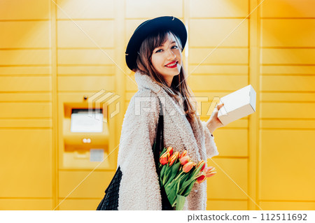 Portrait of smiling fashion woman with box near modern postal automatic mail terminal with self service device for pickup or refund an order. Electronic locker for storing parcels. Selective focus. 112511692