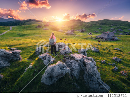 Girl on the stone, cows on green meadows in mountains at sunset Girl on the stone, cows on green meadows in mountains at sunset 112512711