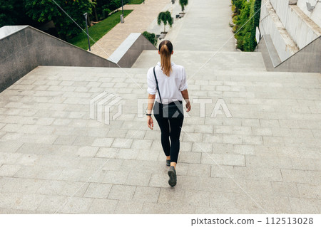 A woman wearing a white shirt and black pants walks up a set of stairs. She is carrying a black purse and a handbag 112513028