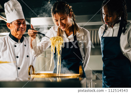 Chef educates in kitchen. Schoolgirls make Japanese noodle. Kids and teacher at stove. Smiling portrait of learning is modern education. Making dinner with ladle gives joy. Foor Education Concept 112514543