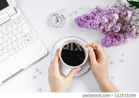 Female hands holding cup of espresso coffee. Alarm clock and lilac flowers on light background. Female hands holding cup of espresso coffee. Alarm clock and lilac flowers on light background. 112515209