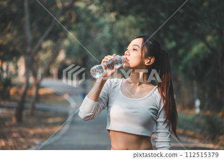 A health-conscious woman takes a refreshing break to drink water during her outdoor exercise routine, highlighting the importance of hydration in fitness. 112515379