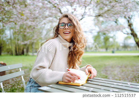 Young woman sitting at a table in blooming spring park with a notepad and laptop. Lifestyle concept. 112515537