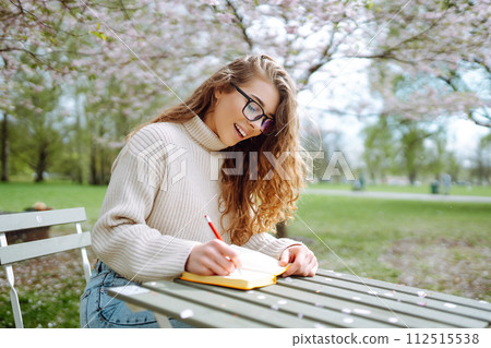 Young woman sitting at a table in blooming spring park with a notepad and laptop. Lifestyle concept. Young woman sitting at a table in blooming spring park with a notepad and laptop. Lifestyle concept. 112515538