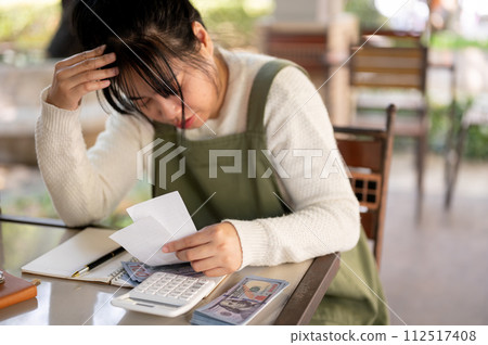 A stressed Asian female small business owner is calculating bills, worried about her shop expenses. A stressed Asian female small business owner is calculating bills, worried about her shop expenses. 112517408