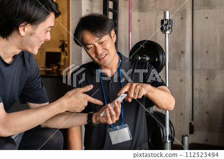 A male trainer and a male member use smartphones to sign up and provide guidance at a sports gym/personal gym. A male trainer and a male member use smartphones to sign up and provide guidance at a sports gym/personal gym. 112523494