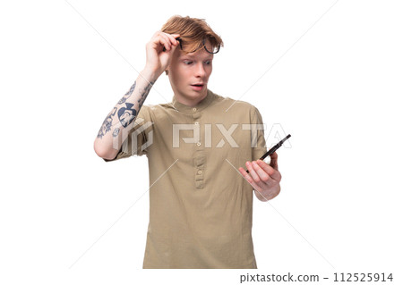 young handsome caucasian guy with red hair in a brown shirt looks at the phone taking off his young handsome caucasian guy with red hair in a brown shirt looks at the phone taking off his 112525914