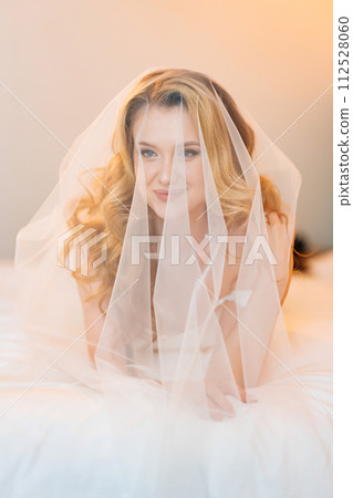 Morning of the bride. Delicate and beautiful blonde bride in bed under veil. 112528060