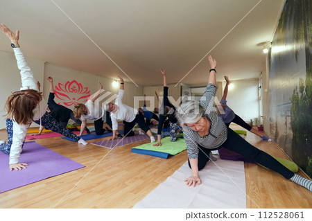 A group of senior women engage in various yoga exercises, including neck, back, and leg stretches, under the guidance of a trainer in a sunlit space, promoting well-being and harmony 112528061