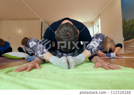 A group of senior women engage in various yoga exercises, including neck, back, and leg stretches, under the guidance of a trainer in a sunlit space, promoting well-being and harmony A group of senior women engage in various yoga exercises, including neck, back, and leg stretches, under the guidance of a trainer in a sunlit space, promoting well-being and harmony 112528188