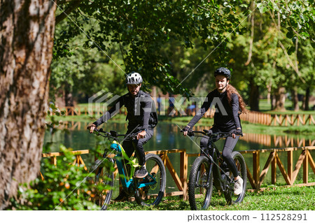 A blissful couple, adorned in professional cycling gear, enjoys a romantic bicycle ride through a park, surrounded by modern natural attractions, radiating love and happiness A blissful couple, adorned in professional cycling gear, enjoys a romantic bicycle ride through a park, surrounded by modern natural attractions, radiating love and happiness 112528291