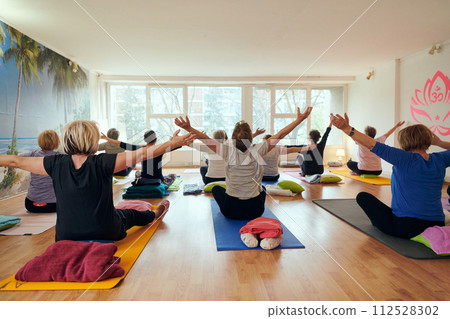A group of senior women engage in various yoga exercises, including neck, back, and leg stretches, under the guidance of a trainer in a sunlit space, promoting well-being and harmony 112528302