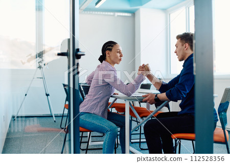 In a modern startup office, a businessman and a businesswoman business colleagues engage in a symbolic arm-wrestling match, reflecting teamwork, competition, and innovation in their dynamic workplace. In a modern startup office, a businessman and a businesswoman business colleagues engage in a symbolic arm-wrestling match, reflecting teamwork, competition, and innovation in their dynamic workplace. 112528356