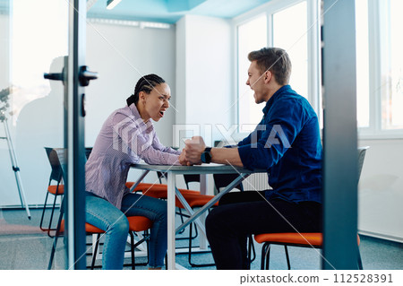 In a modern startup office, a businessman and a businesswoman business colleagues engage in a symbolic arm-wrestling match, reflecting teamwork, competition, and innovation in their dynamic workplace. In a modern startup office, a businessman and a businesswoman business colleagues engage in a symbolic arm-wrestling match, reflecting teamwork, competition, and innovation in their dynamic workplace. 112528391