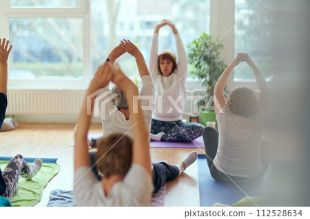 A group of senior women engage in various yoga exercises, including neck, back, and leg stretches, under the guidance of a trainer in a sunlit space, promoting well-being and harmony 112528634