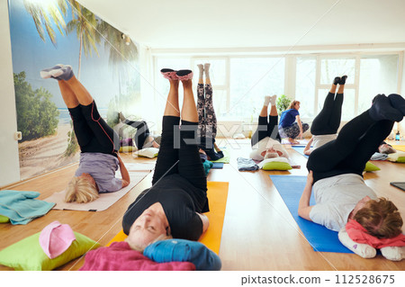 A group of senior women engage in various yoga exercises, including neck, back, and leg stretches, under the guidance of a trainer in a sunlit space, promoting well-being and harmony 112528675