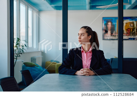 Successful young female leader in a suit with a pink shirt sitting in a modern glass office with a determined smile. 112528684