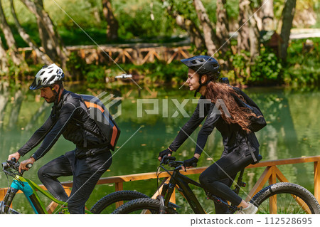A blissful couple, adorned in professional cycling gear, enjoys a romantic bicycle ride through a park, surrounded by modern natural attractions, radiating love and happiness 112528695