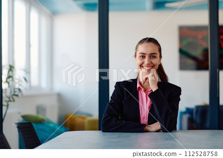 Successful young female leader in a suit with a pink shirt sitting in a modern glass office with a determined smile. 112528758