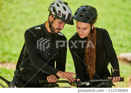 A blissful couple, adorned in professional cycling gear, enjoys a romantic bicycle ride through a park, surrounded by modern natural attractions, radiating love and happiness 112530064