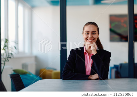 Successful young female leader in a suit with a pink shirt sitting in a modern glass office with a determined smile. Successful young female leader in a suit with a pink shirt sitting in a modern glass office with a determined smile. 112530065