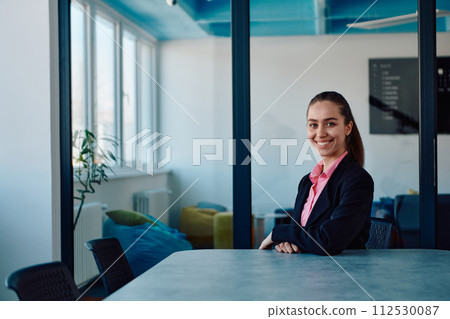 Successful young female leader in a suit with a pink shirt sitting in a modern glass office with a determined smile. 112530087