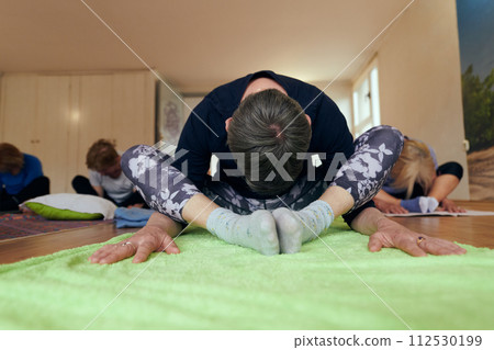 A group of senior women engage in various yoga exercises, including neck, back, and leg stretches, under the guidance of a trainer in a sunlit space, promoting well-being and harmony 112530199