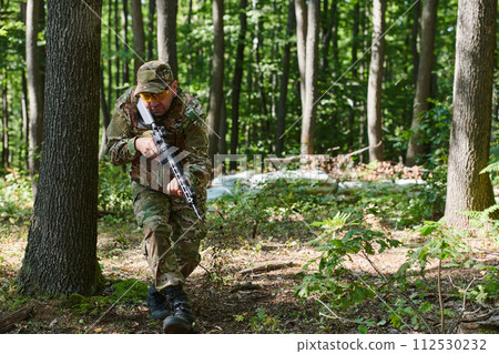 An elite soldier, camouflaged and stealthily navigating through dangerous woodland terrain, executes a covert mission in a secluded forest area An elite soldier, camouflaged and stealthily navigating through dangerous woodland terrain, executes a covert mission in a secluded forest area 112530232