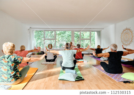 A group of senior women engage in various yoga exercises, including neck, back, and leg stretches, under the guidance of a trainer in a sunlit space, promoting well-being and harmony 112530315