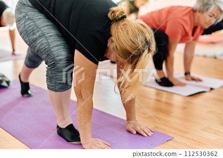 A group of senior women engage in various yoga exercises, including neck, back, and leg stretches, under the guidance of a trainer in a sunlit space, promoting well-being and harmony 112530362