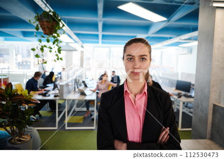 Portrait of young smiling business woman in creative open space coworking startup office. Successful businesswoman standing in office with copyspace. Coworkers working in background Portrait of young smiling business woman in creative open space coworking startup office. Successful businesswoman standing in office with copyspace. Coworkers working in background 112530473