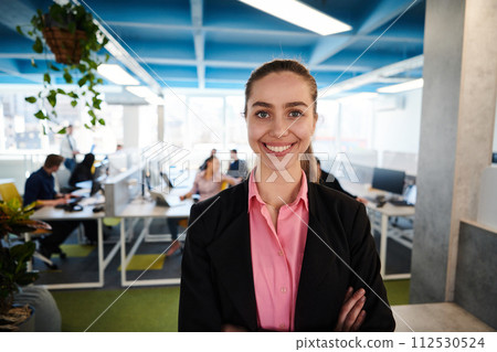 Portrait of young smiling business woman in creative open space coworking startup office. Successful businesswoman standing in office with copyspace. Coworkers working in background Portrait of young smiling business woman in creative open space coworking startup office. Successful businesswoman standing in office with copyspace. Coworkers working in background 112530524