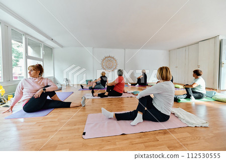 A group of senior women engage in various yoga exercises, including neck, back, and leg stretches, under the guidance of a trainer in a sunlit space, promoting well-being and harmony 112530555