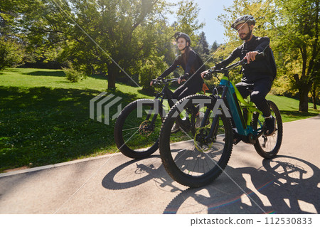 A blissful couple, adorned in professional cycling gear, enjoys a romantic bicycle ride through a park, surrounded by modern natural attractions, radiating love and happiness 112530833