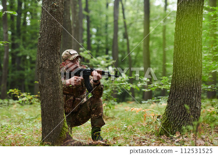 An elite soldier, camouflaged and stealthily navigating through dangerous woodland terrain, executes a covert mission in a secluded forest area 112531525