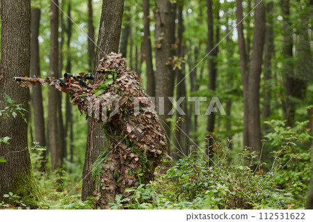 A highly skilled elite sniper, camouflaged in the dense forest, stealthily maneuvers through dangerous woodland terrain on a covert and precise mission 112531622